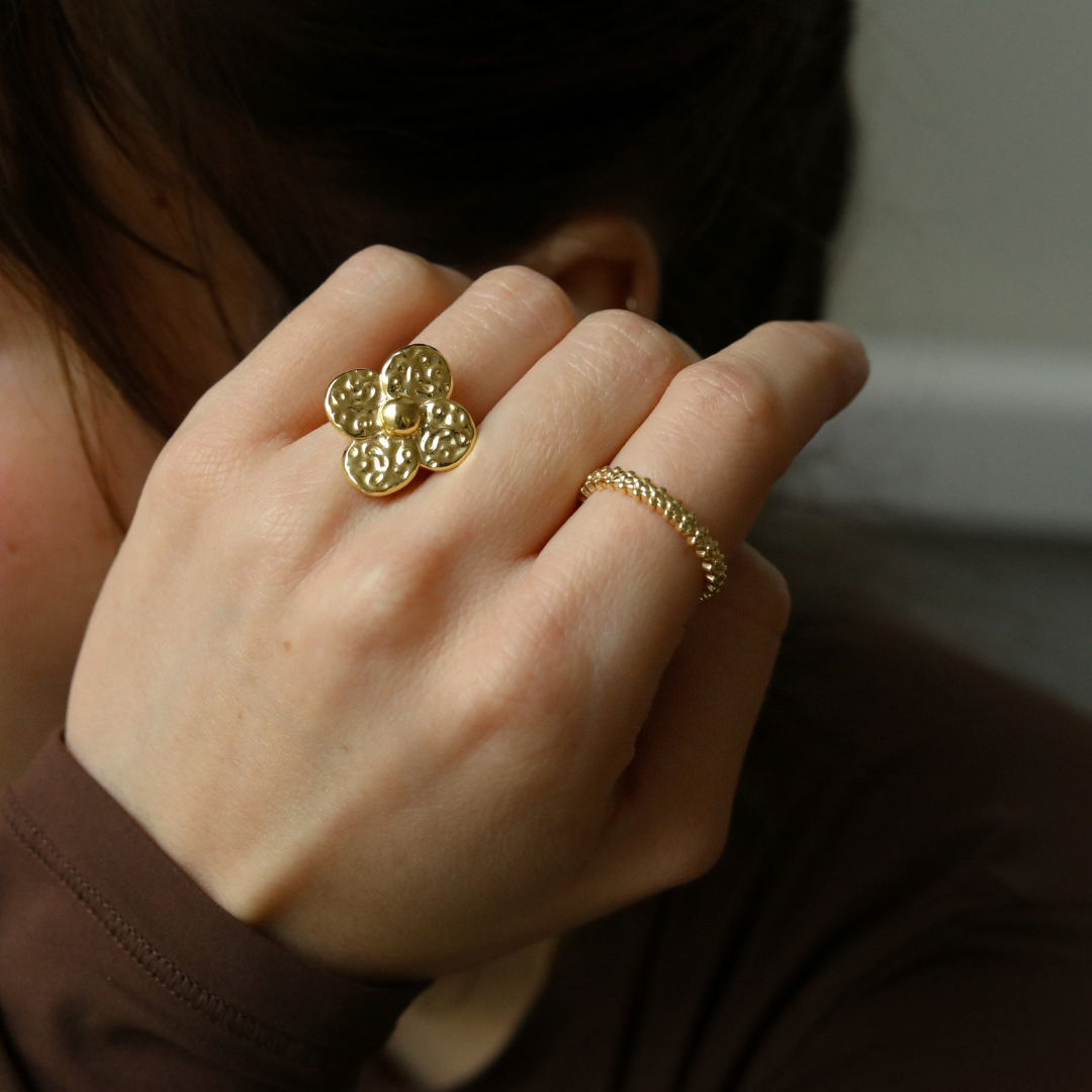 Hand met goudkleurige bloem statement ring, gecombineerd met een fijn gouden ringetje – opvallend en stijlvol accessoire.